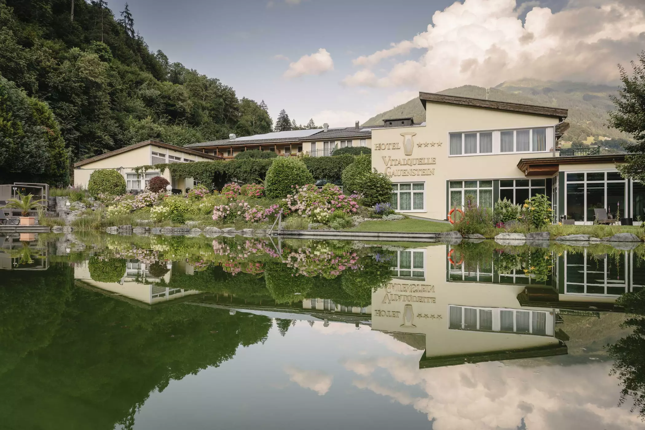 Hotelgebäude mit Garten und blühenden Hortensien am Naturteich, in dem sich die Fassade und die Berge spiegeln.