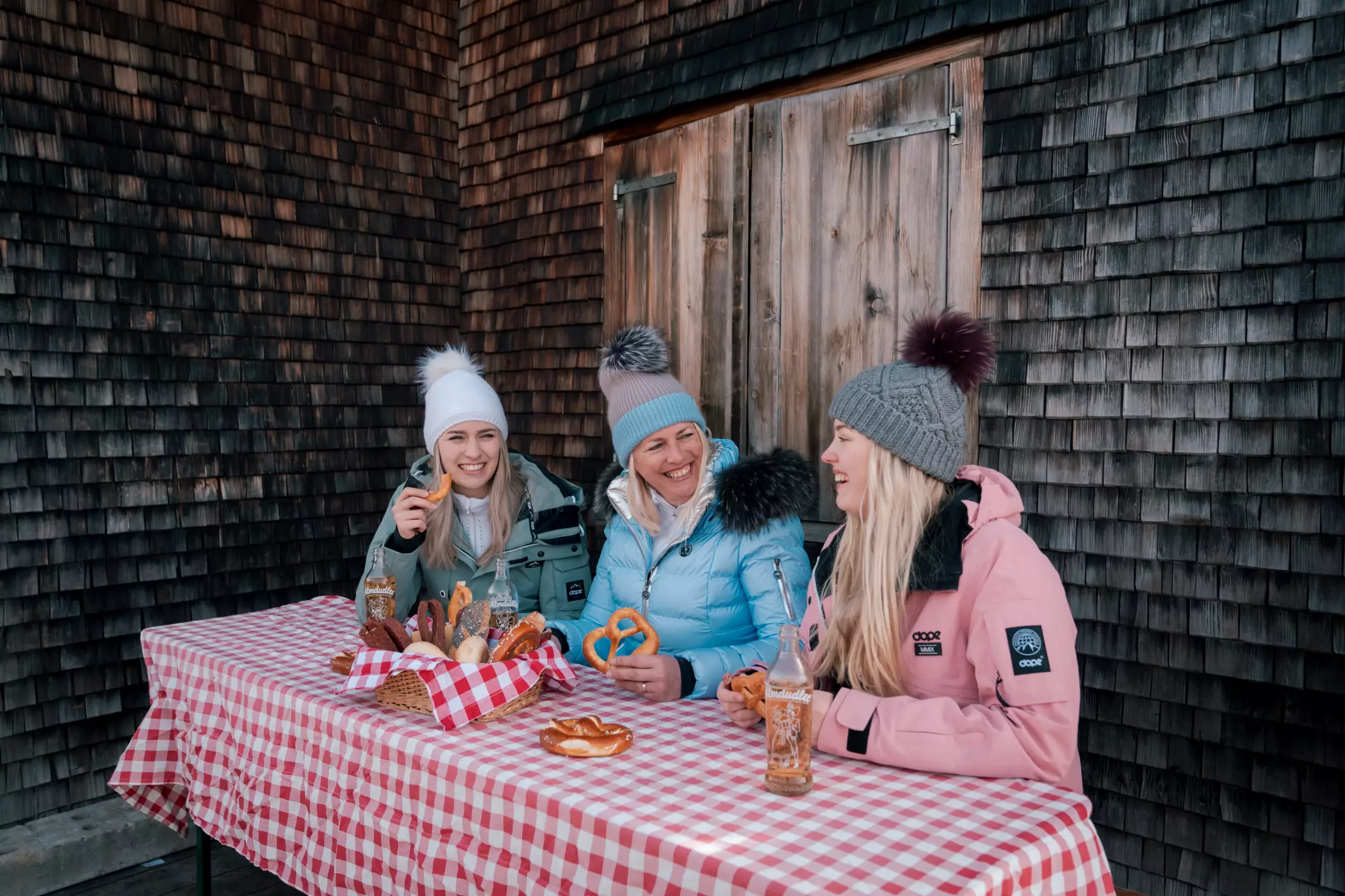 Drei fröhliche Frauen in warmer Winterkleidung sitzen draußen an einem rustikalen Holztisch mit rot-weiß karierter Tischdecke. Sie essen bayerische Spezialitäten wie Brezeln und Semmeln und trinken Limonade, vor einem Holzhaus mit Schindelfassade.