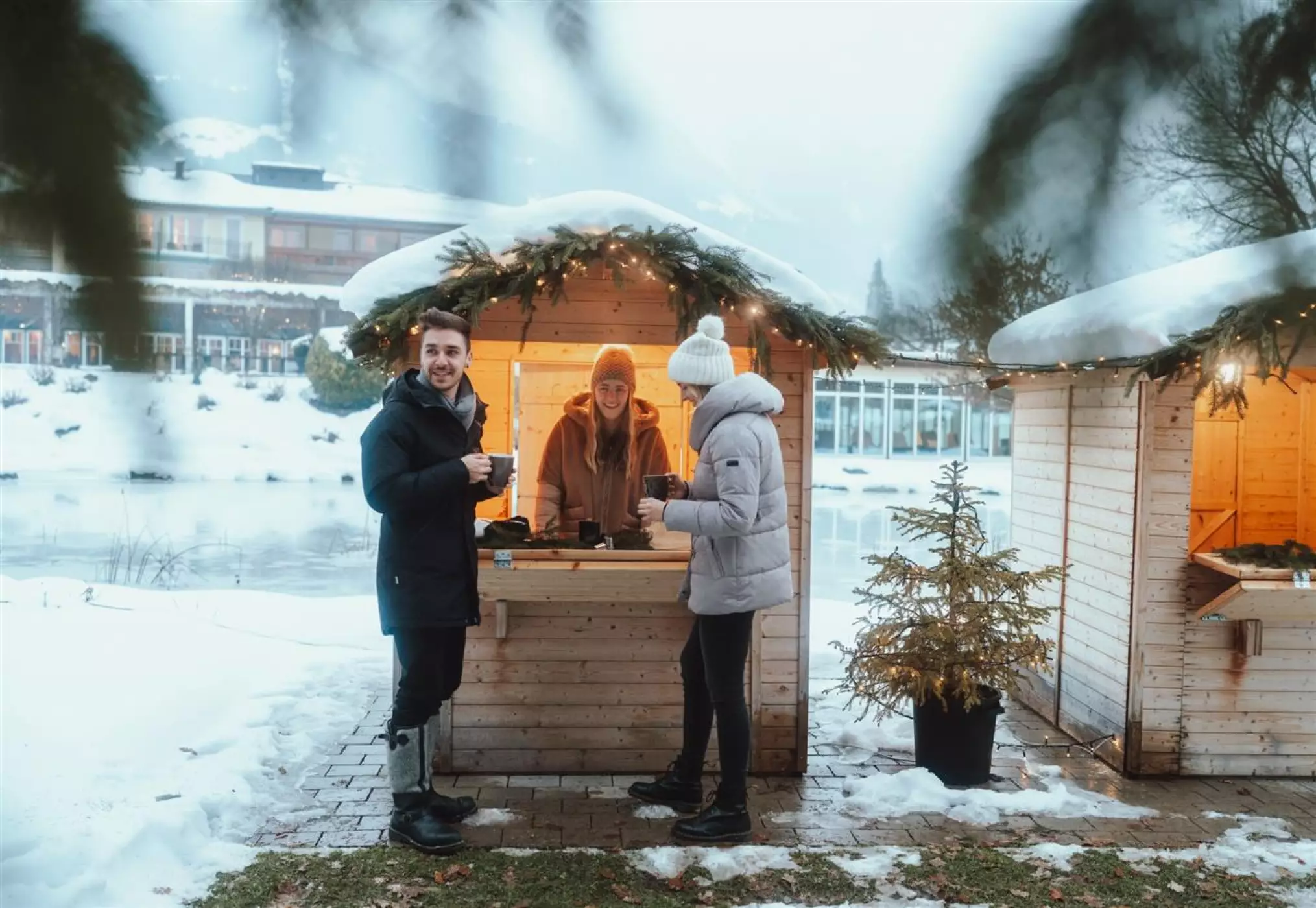 Ein Mann und eine Frau in Winterkleidung genießen heiße Getränke an einem kleinen, festlich dekorierten Weihnachtsstand aus Holz, der von einer freundlich lächelnden Verkäuferin bedient wird. Umgeben von Schnee und mit Blick auf einen zugefrorenen See entsteht eine gemütliche Winterstimmung.