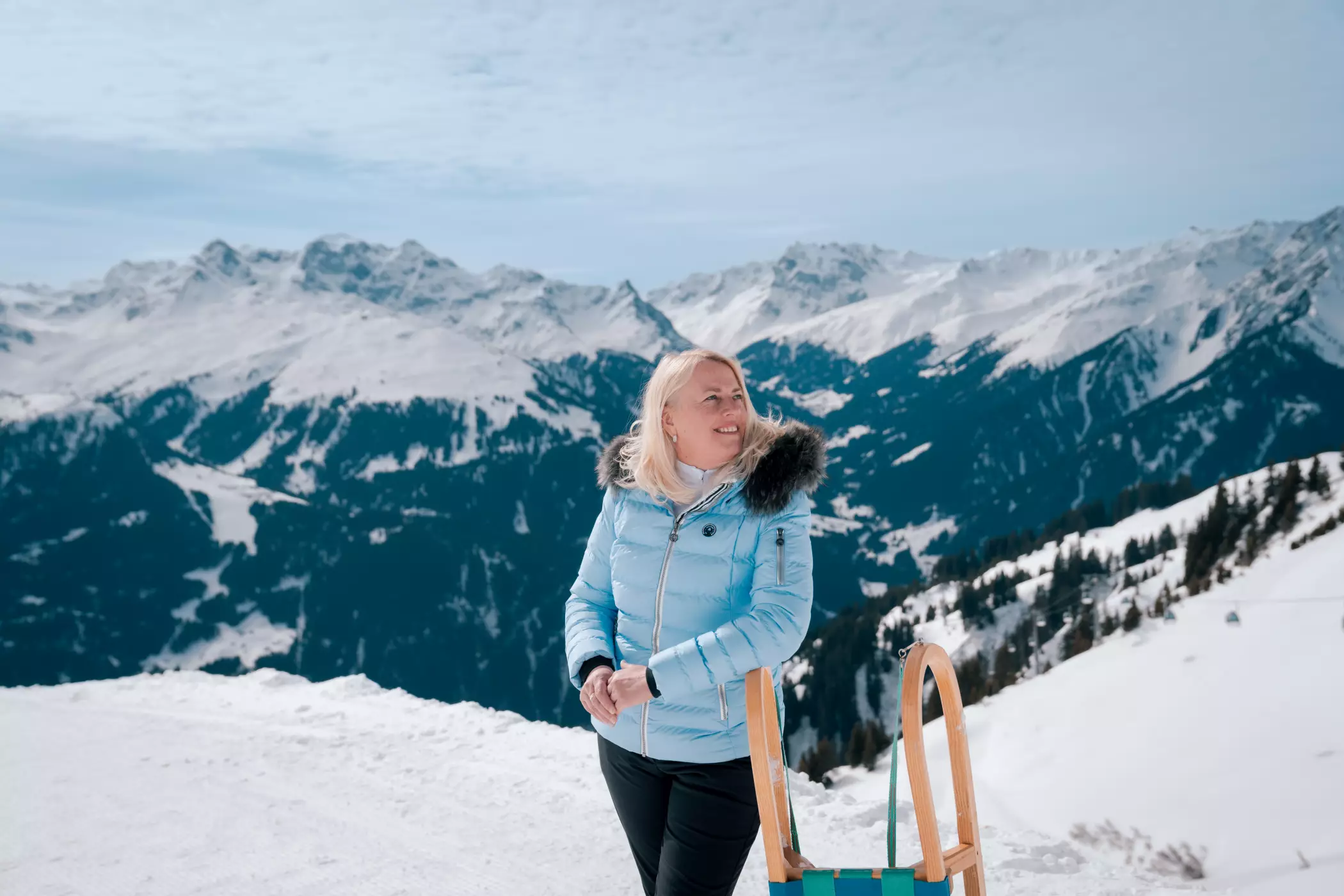 Eine Frau in einer hellblauen Daunenjacke steht mit einem traditionellen Holzschlitten vor einer beeindruckenden Bergkulisse in den verschneiten Alpen. Der sonnige Wintertag und die weite Aussicht über die verschneiten Gipfel erzeugen eine entspannte, naturnahe Stimmung.