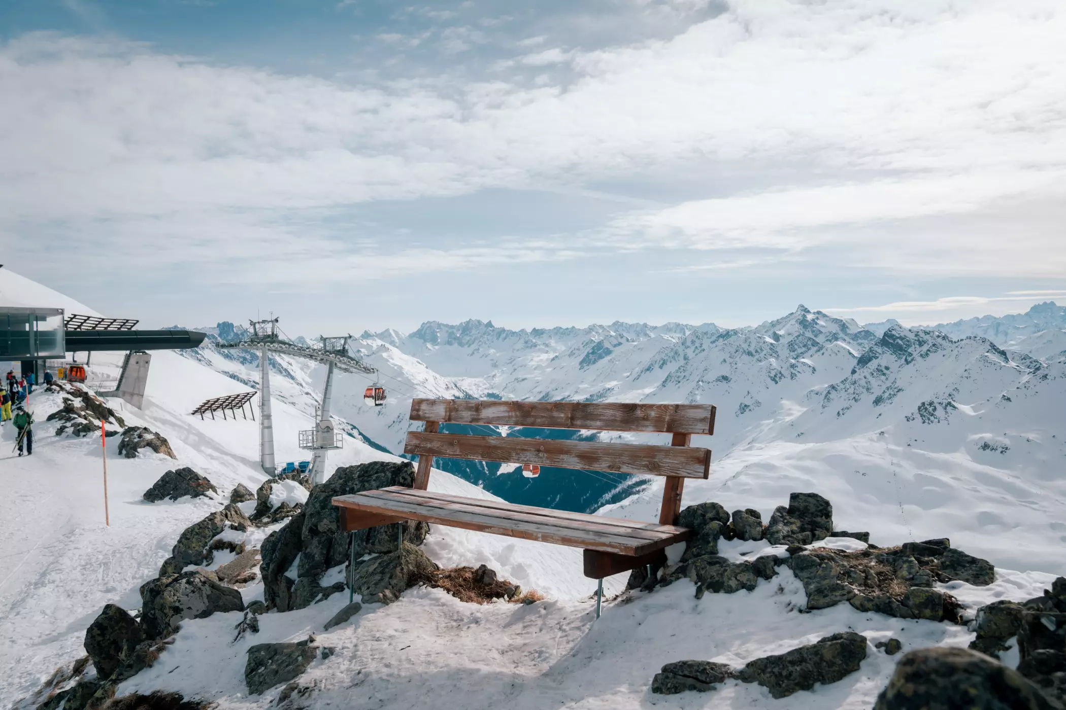 Eine hölzerne Bank steht auf einem schneebedeckten Aussichtspunkt mit Blick auf eine majestätische Berglandschaft in den Alpen. Im Hintergrund sind Sessellifte, ein Skigebiet und schneebedeckte Gipfel unter einem teils bewölkten Himmel zu sehen.